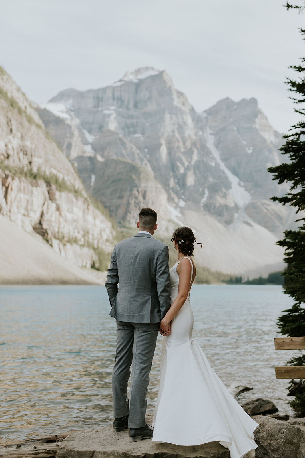 A bride and groom stand hand in hand on a lakeshore in Banff, facing dramatic snow-capped mountains and a calm blue lake—a breathtaking wedding scene with the bride in white and the groom in a gray suit.