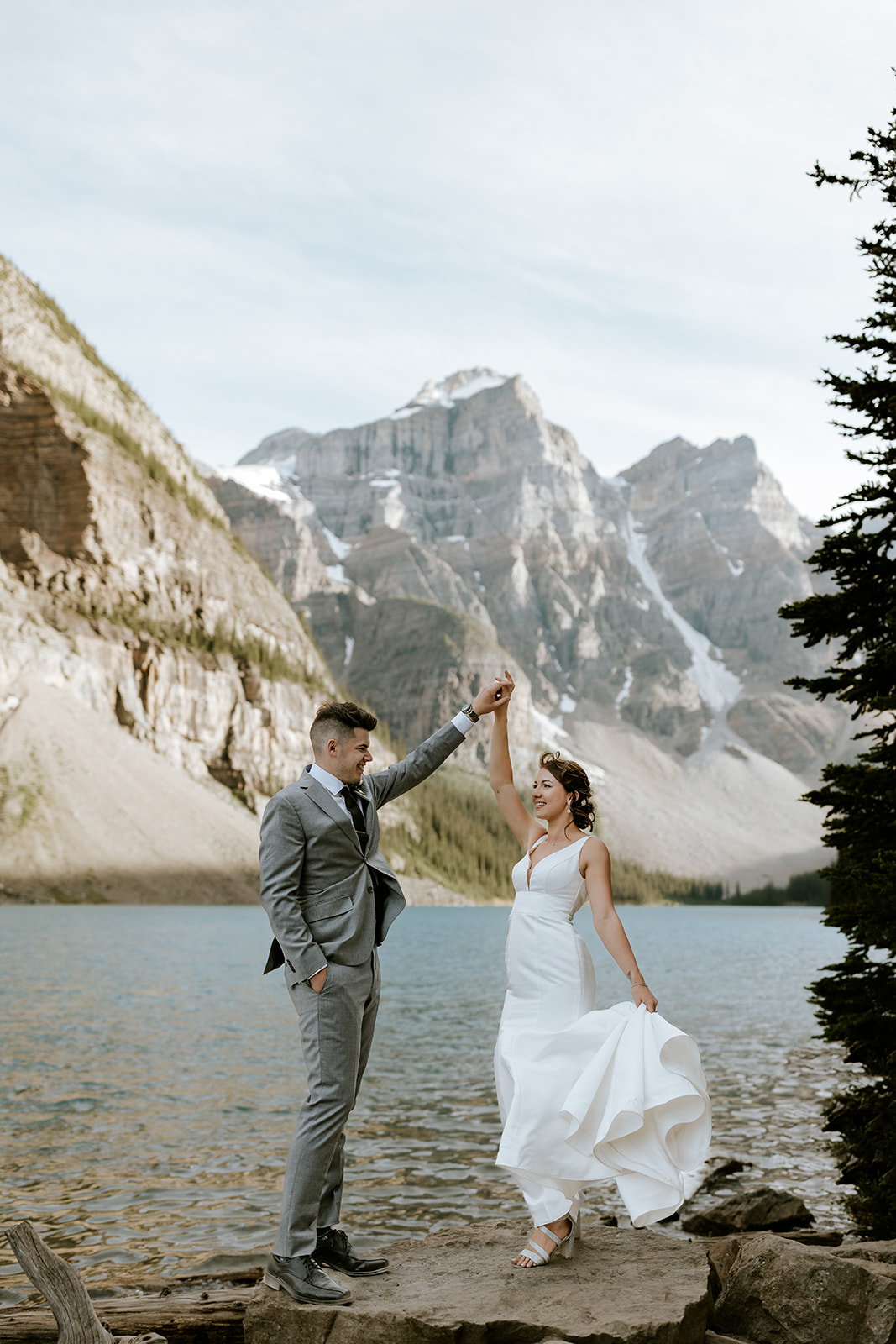 A bride in a white dress and a groom in a gray suit dance joyfully by a mountain lake in Banff, with rocky peaks and evergreens behind them, as their wedding photographer captures the moment beneath a lightly cloudy sky.