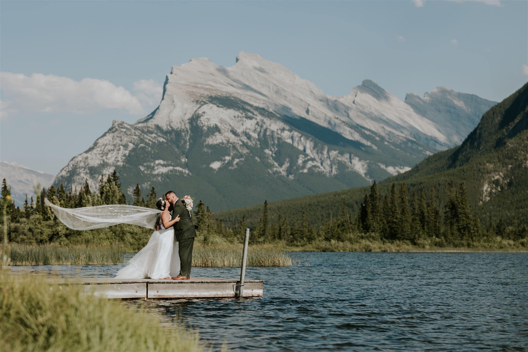 A bride and groom share a kiss on a wooden dock by a lake in Canmore, surrounded by trees and mountains. The bride’s veil flows in the wind as the dramatic peaks create a stunning elopement backdrop.
