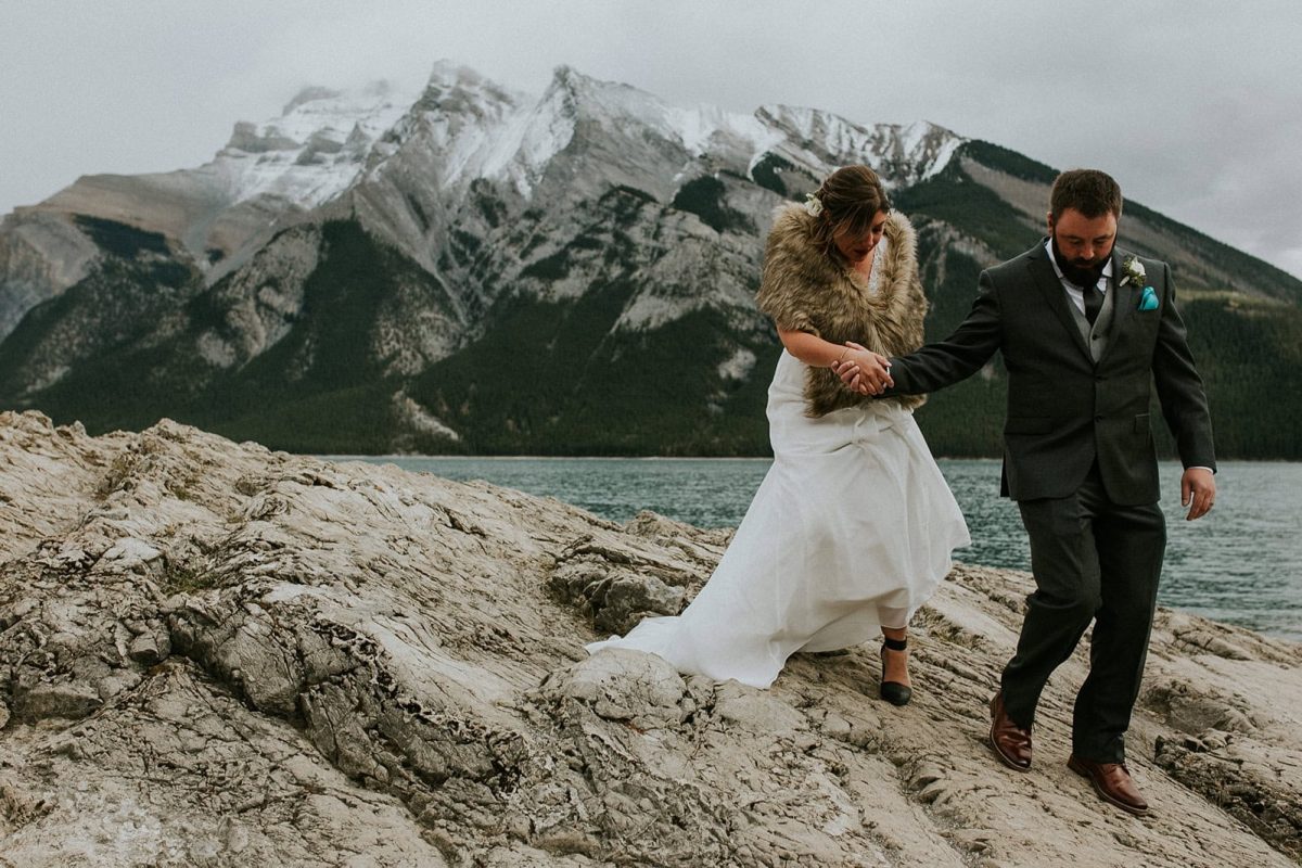 A bride in a white dress and fur shawl and a groom in a dark suit walk on rocky terrain by a lake in Banff, with snow-capped mountains in the background—a picturesque Calgary wedding under a cloudy sky.