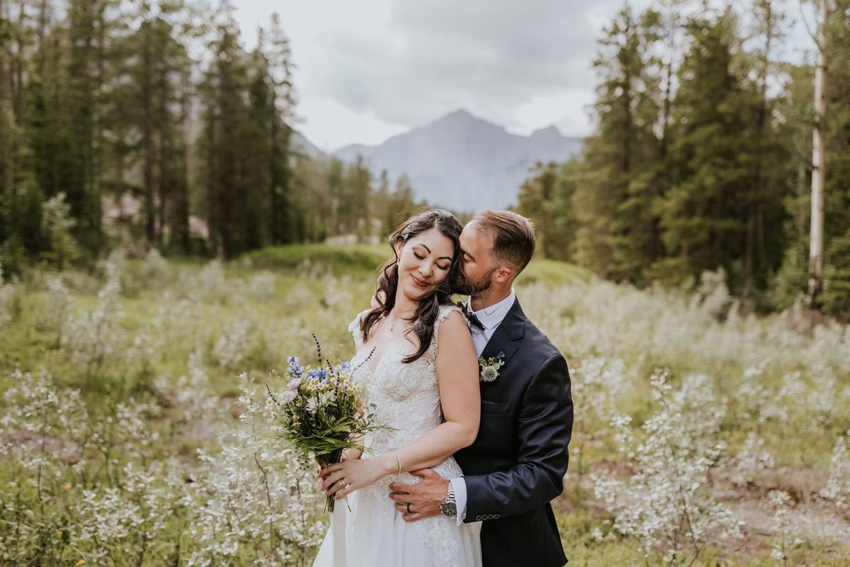 A bride and groom embrace in a field with wildflowers and tall trees near Calgary, mountains in the background. The bride holds a bouquet and smiles with her eyes closed as the wedding photographer captures the groom gently kissing her cheek.