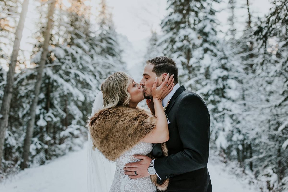 A bride and groom share a kiss outdoors in a snowy forest near Calgary. The bride wears a white dress, veil, and fur stole, while the groom is in a dark suit. Snow-covered trees surround them, captured perfectly by their wedding photographer.