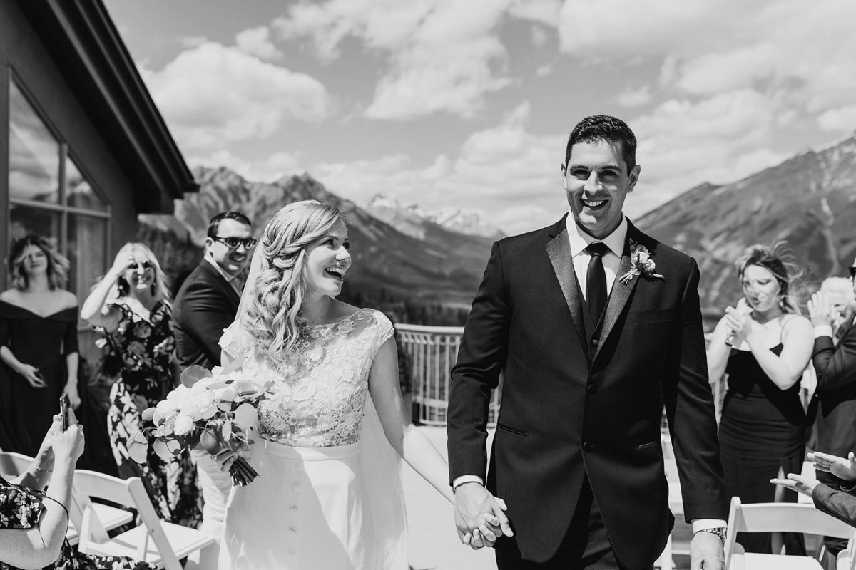 A bride and groom smile joyfully while holding hands and walking down the aisle outdoors in Canmore, with mountains in the background as guests clap and a photographer captures every moment.