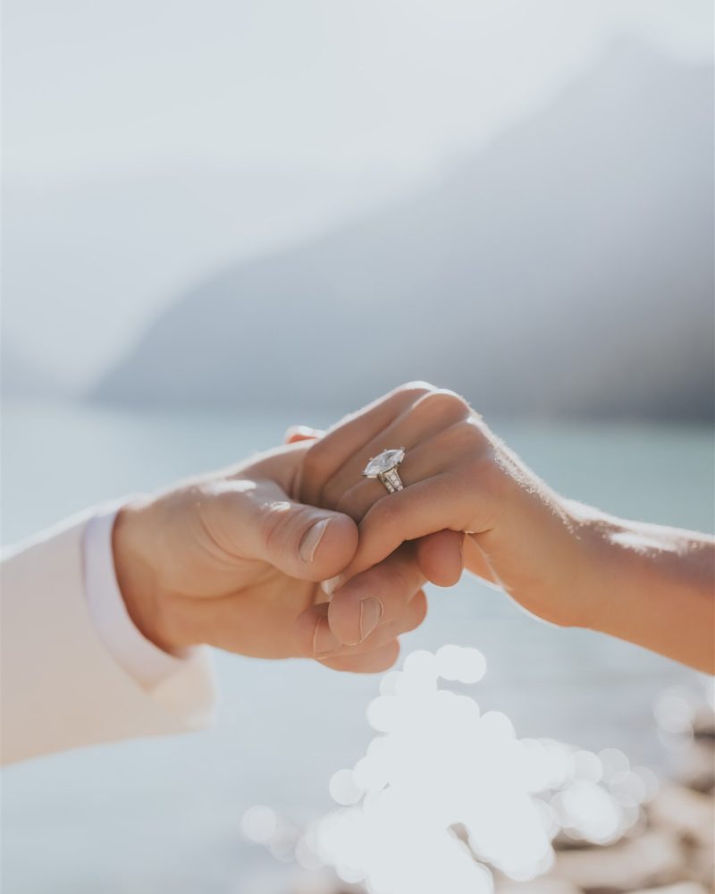 Close-up of two hands holding each other, one with a diamond engagement ring. Captured by a Calgary elopement photographer, the blurred background features sunlight shimmering on water and distant mountains.
