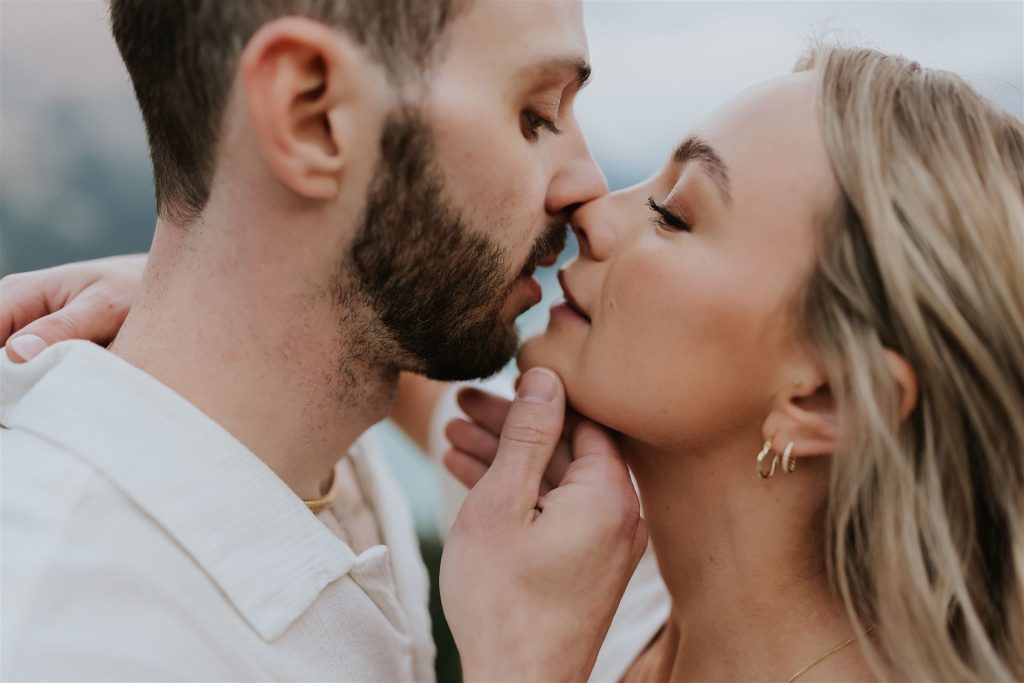 A close-up of a couple about to kiss, with the man gently holding the woman's chin. Both have their eyes closed and appear affectionate, set against a softly blurred outdoor Banff background—perfect for an intimate elopement.
