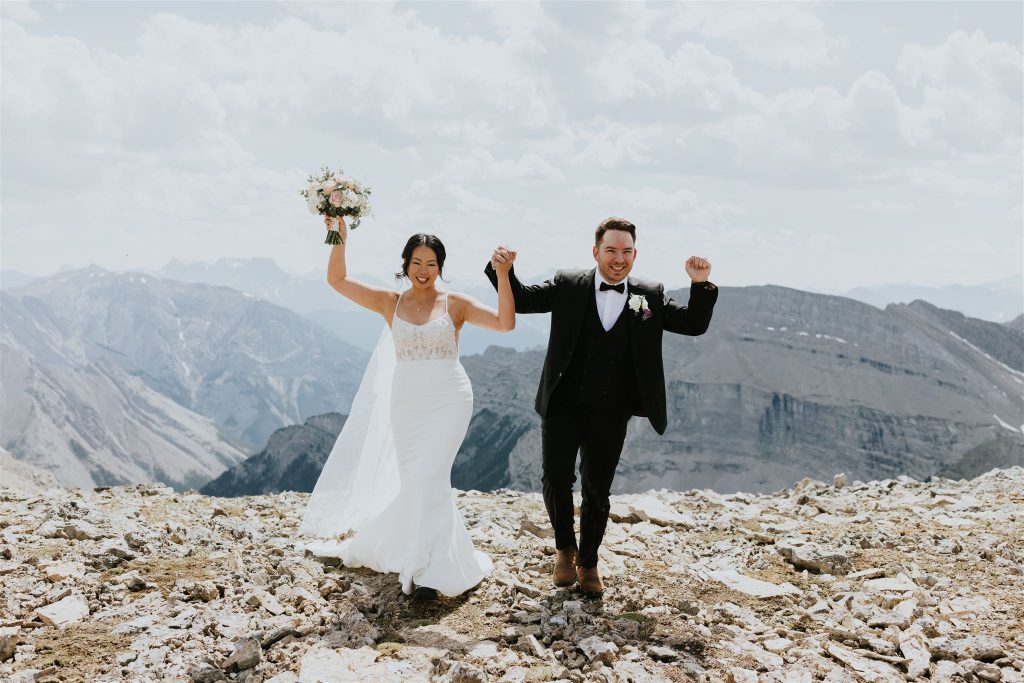 A bride and groom celebrate on a rocky mountaintop in Banff, holding hands and raising their arms joyfully, with mountains and a cloudy sky in the background—captured by a local photographer.