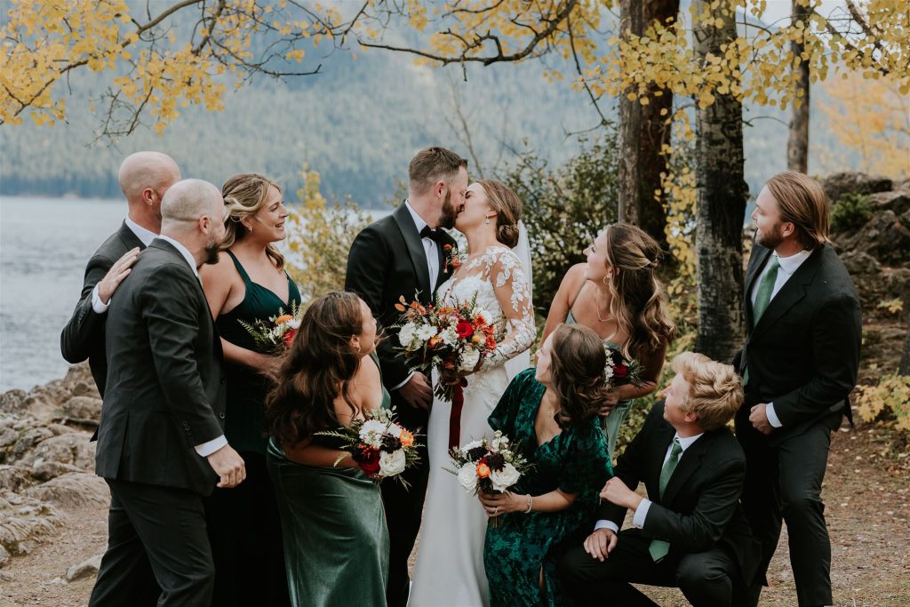 A bride and groom kiss outdoors by a lake near Canmore, surrounded by their joyful wedding party in dark green and black attire, holding bouquets, with autumn trees and mountains in the background.