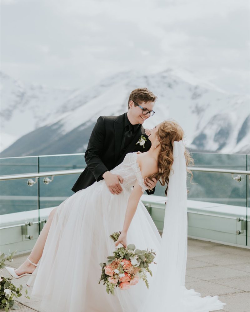 A groom in a black suit dips his bride in a white wedding dress and veil on a balcony, with snow-capped Banff mountains in the background. The bride holds a bouquet of flowers and they are smiling at each other on their wedding day.