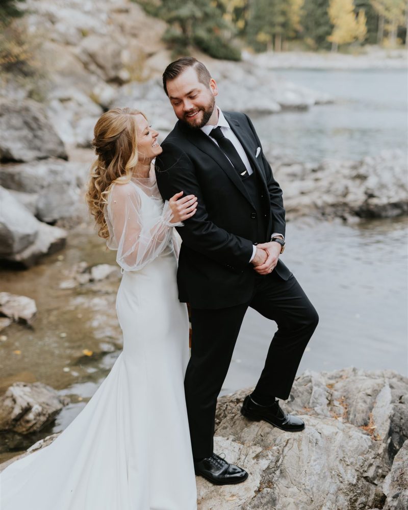 A bride in a white dress and a groom in a black suit stand on rocks by a lakeshore, smiling and laughing together with trees and water in the background—captured by a Calgary photographer during their intimate elopement.