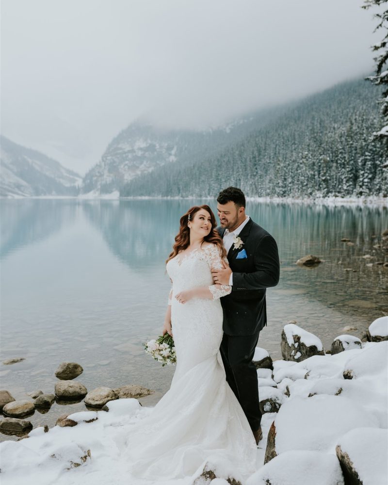 A bride in a white gown and a groom in a black suit stand on snowy rocks by a calm, turquoise lake in Banff, surrounded by snowy mountains and evergreen trees, embracing each other as their wedding photographer captures the moment.