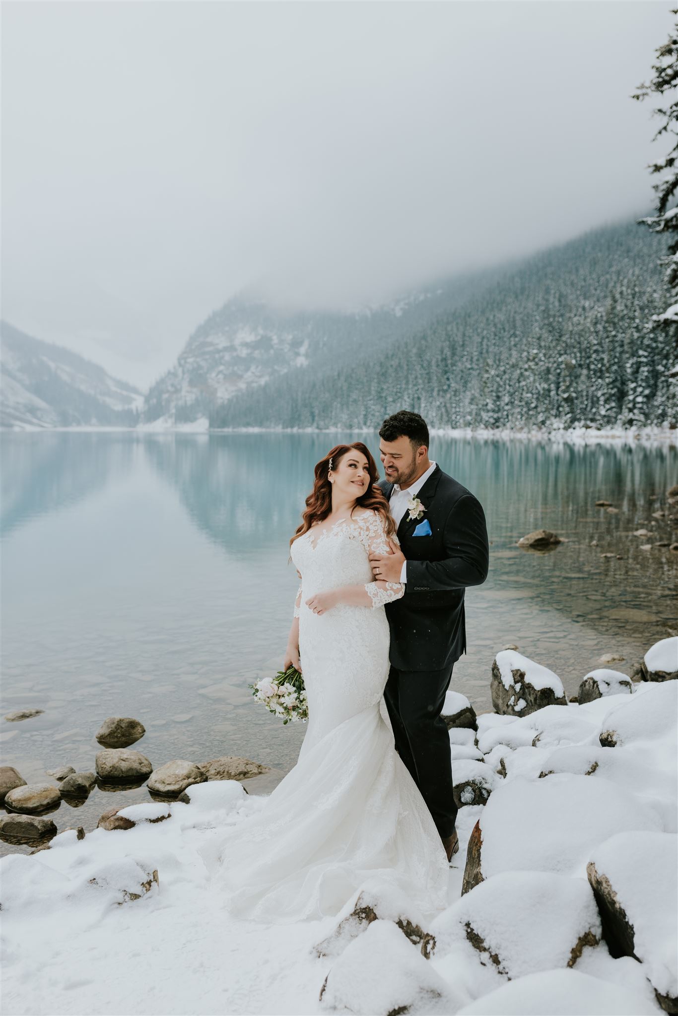 A bride in a white gown and a groom in a black suit stand on snowy rocks by a calm, turquoise lake in Banff, surrounded by snowy mountains and evergreen trees, embracing each other as their wedding photographer captures the moment.