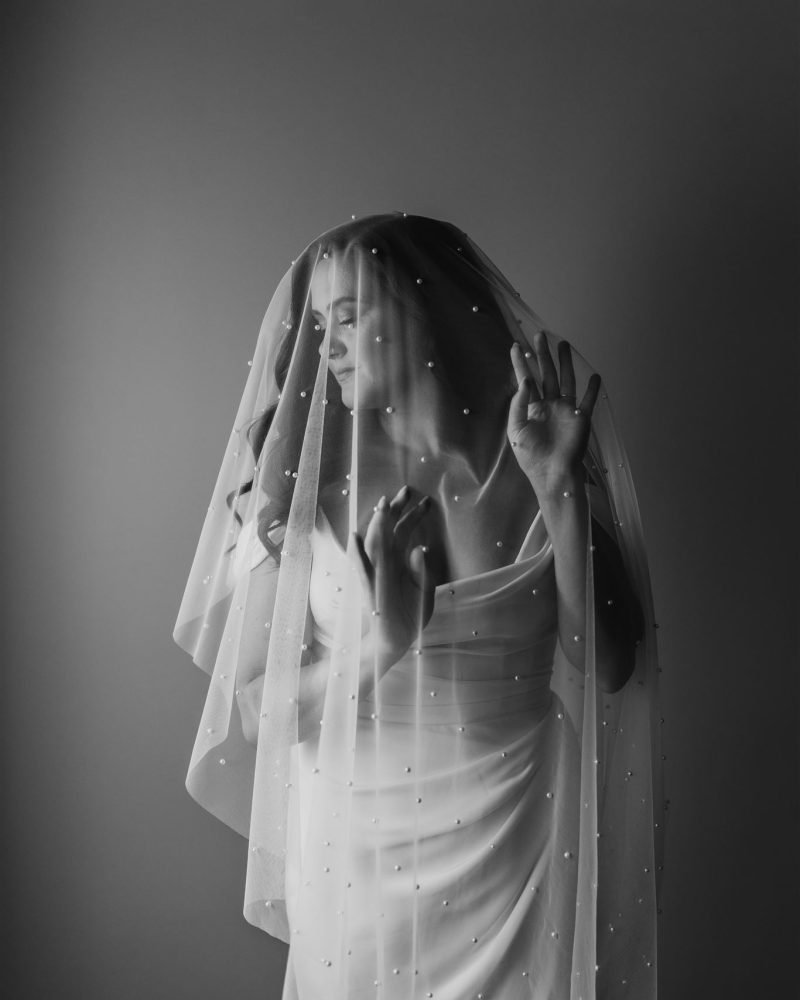 A woman in a white dress stands with her hands raised, draped in a sheer, beaded veil that covers her head and shoulders. Captured by a Calgary photographer, the black and white photo creates a soft, dreamy atmosphere.