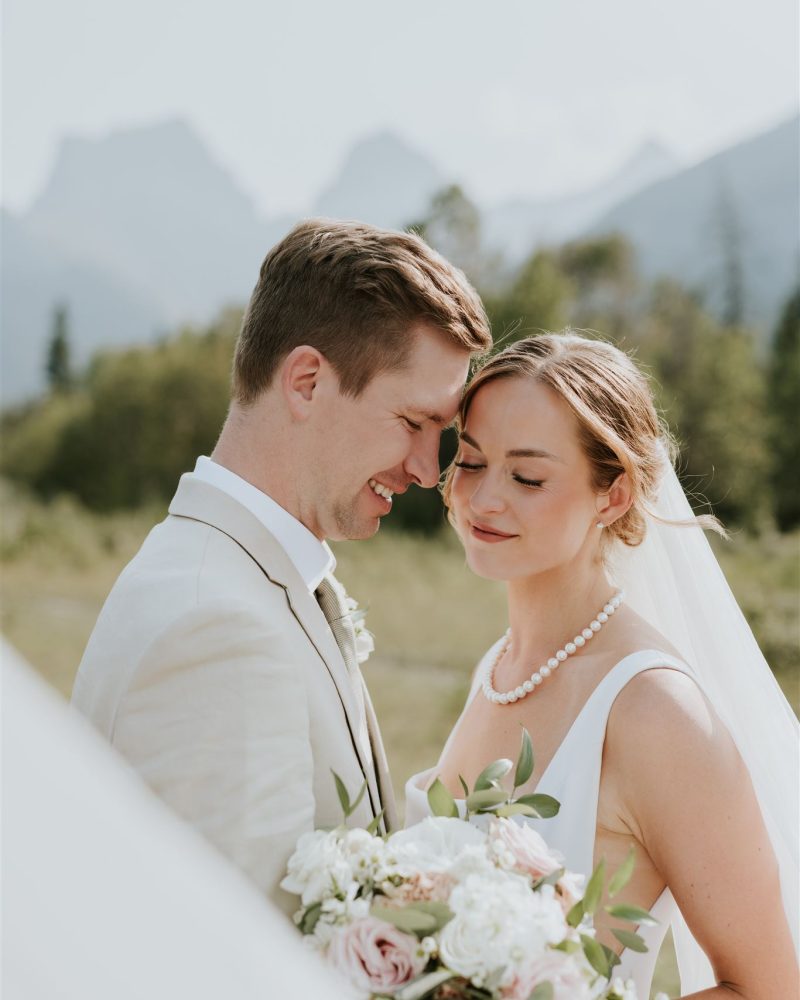 A bride and groom in wedding attire stand closely together outdoors, smiling with their foreheads touching. The bride holds a bouquet of white and pink flowers, with mountains and greenery in the Canmore background captured by a Calgary photographer.