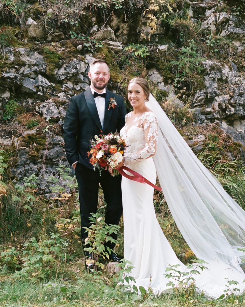 A bride and groom stand outdoors by a rocky hillside with greenery. Captured by a Calgary elopement photographer, the bride in a lace-sleeved white dress and veil holds a bouquet, while the groom in a black suit smiles at the camera.