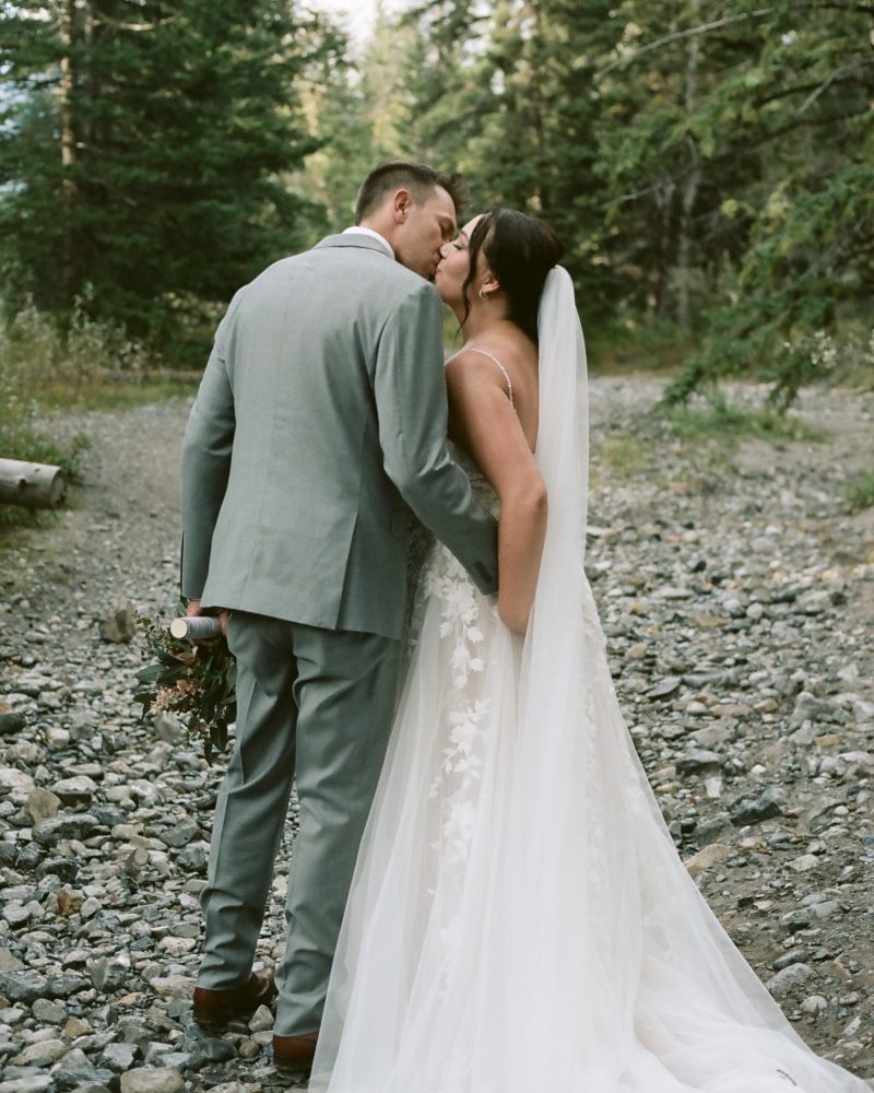 A bride in a flowing white dress and veil kisses her groom in a light gray suit while walking on a rocky forest path, surrounded by tall green trees, capturing a magical Banff wedding moment.
