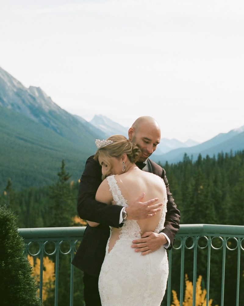 A bride and groom embrace on a balcony overlooking a forest and mountain range near Banff, with snow-capped peaks and trees in the background under a partly cloudy sky—a perfect elopement moment.