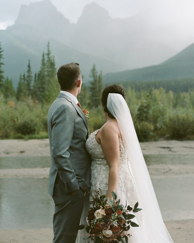 A bride and groom in wedding attire stand side by side, facing a scenic Canmore mountain landscape with pine trees and a river—a perfect setting for an intimate elopement. The bride holds a bouquet and wears a long veil, while the groom wears a gray suit.