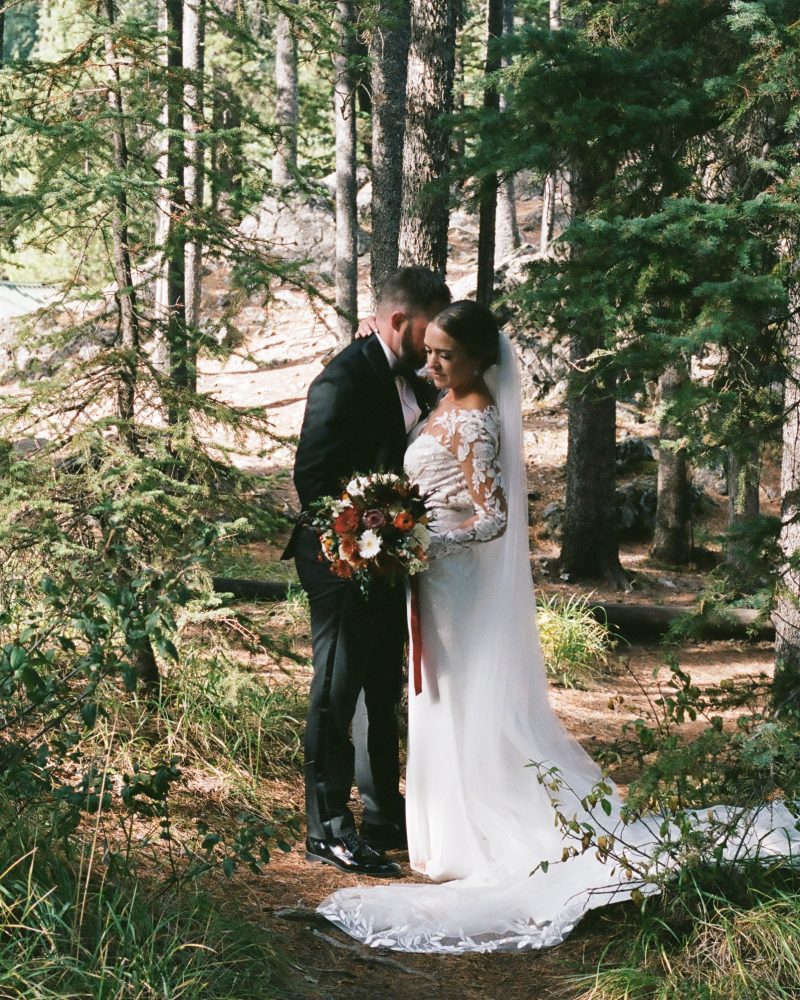A bride and groom stand close together in a forest near Canmore, with the bride in a long white dress holding a bouquet. Sunlight filters through the trees, creating a serene and intimate wedding atmosphere.