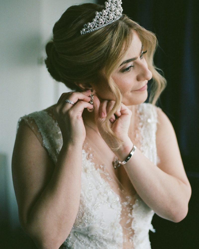 A bride in an embellished white wedding dress and jeweled tiara smiles softly as she puts on a dangling earring, with soft natural light illuminating her face and hair in beautiful Canmore.