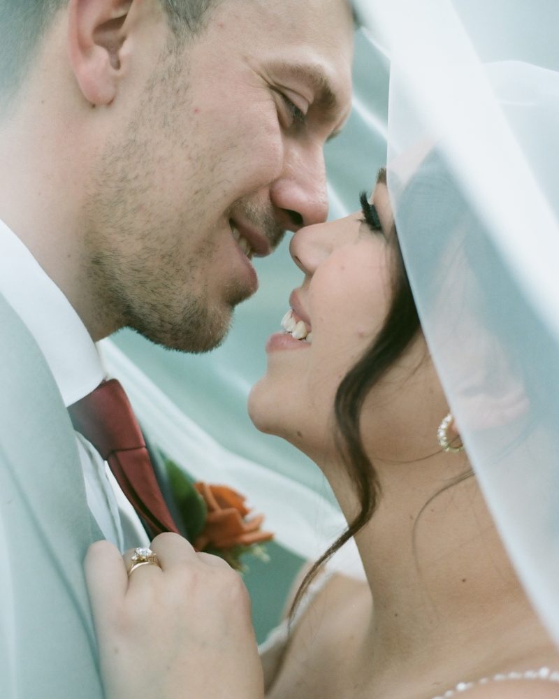 A bride and groom smile lovingly at each other under a white veil, their faces close together. Celebrating their joyful Canmore elopement, the bride touches the groom’s chest as they share an intimate moment on their wedding day.