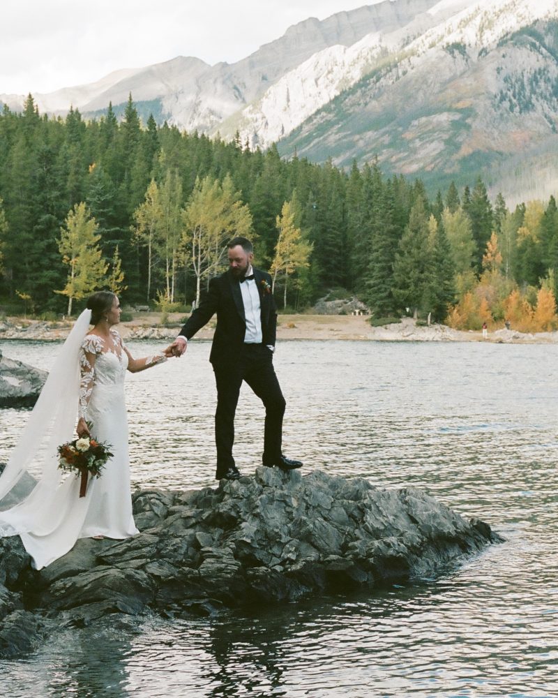 A bride and groom stand on rocky terrain by a lake, holding hands. The bride in a white dress holds a bouquet, while the groom wears a suit. Pine trees and mountains set the scene for their Banff elopement.