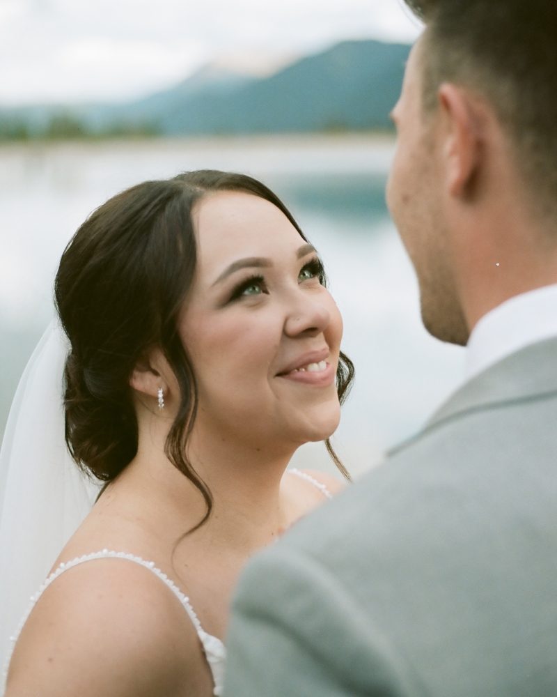 A bride in a white dress and veil smiles lovingly at her groom in a light gray suit, as their Calgary wedding photographer captures the moment against a blurred outdoor backdrop of water and mountains.