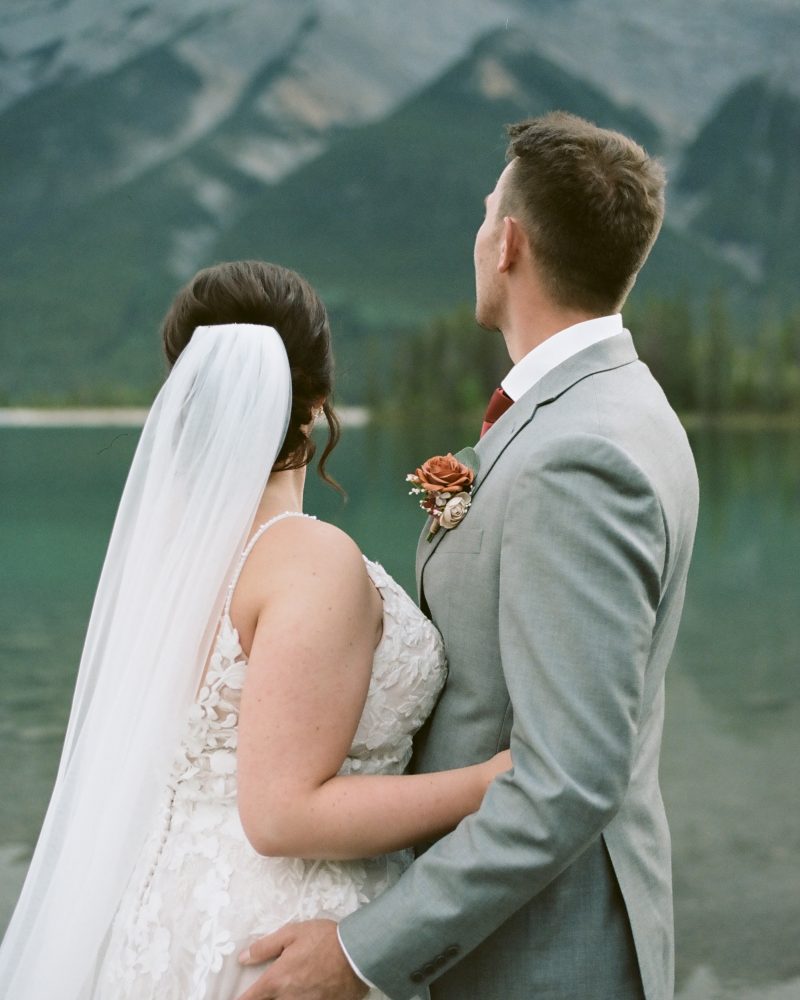 A bride and groom stand arm in arm by a lake in Canmore, facing away and looking at distant mountains. The bride wears a white lace dress and veil; the groom is in a gray suit with a boutonniere, capturing a stunning wedding moment.