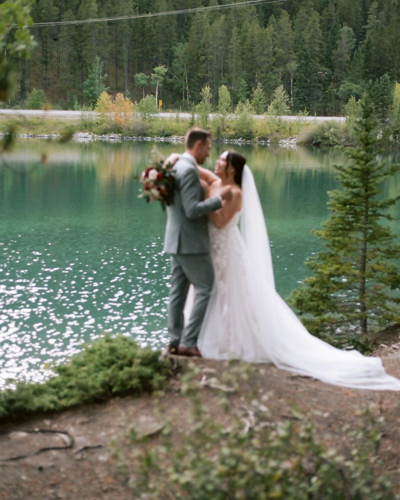 A bride and groom stand close together by a lake in Banff, surrounded by trees. The bride wears a long white dress and veil, while the groom, holding a bouquet, enjoys the stunning water and forest backdrop captured by their wedding photographer.