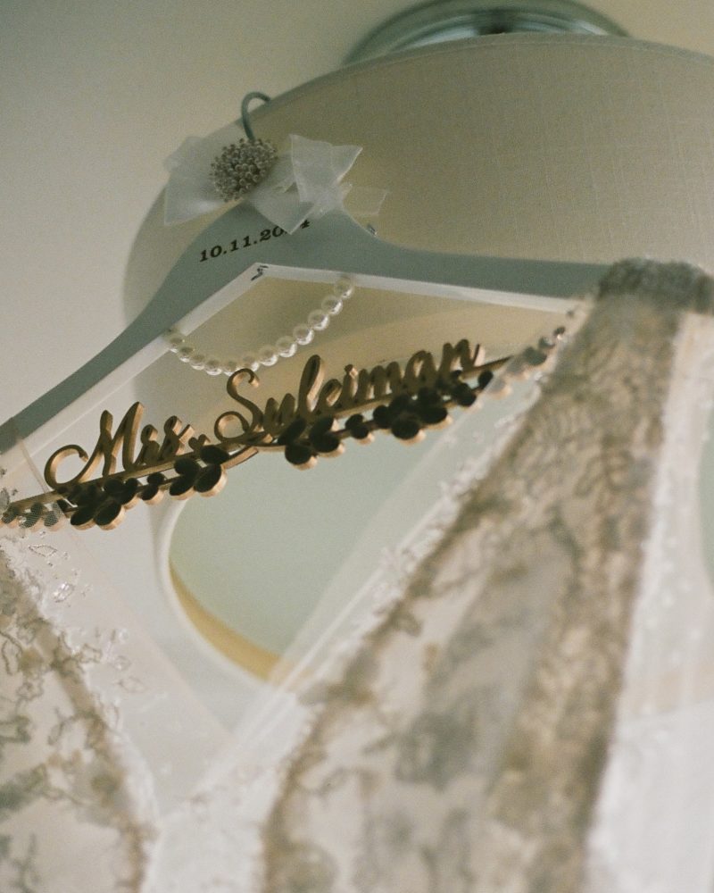 A close-up of a lace wedding dress hanging on a personalized hanger that reads “Mrs. Sullivan.” Ideal for a Canmore elopement, the pearl-adorned hanger with date tag is set in front of a beige lampshade.