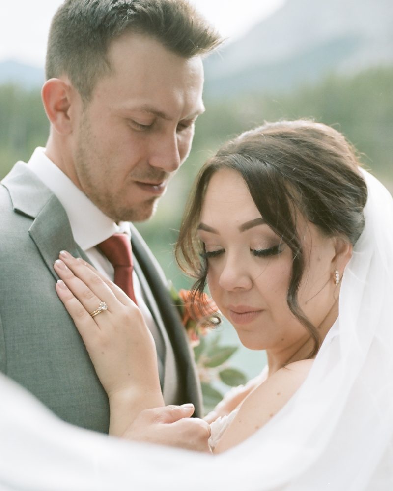 A bride and groom pose closely outdoors during their Banff elopement. The bride, eyes closed with a veil draped around her, rests her hand on the groom’s chest as he gently looks down. Natural scenery is beautifully blurred in the background.