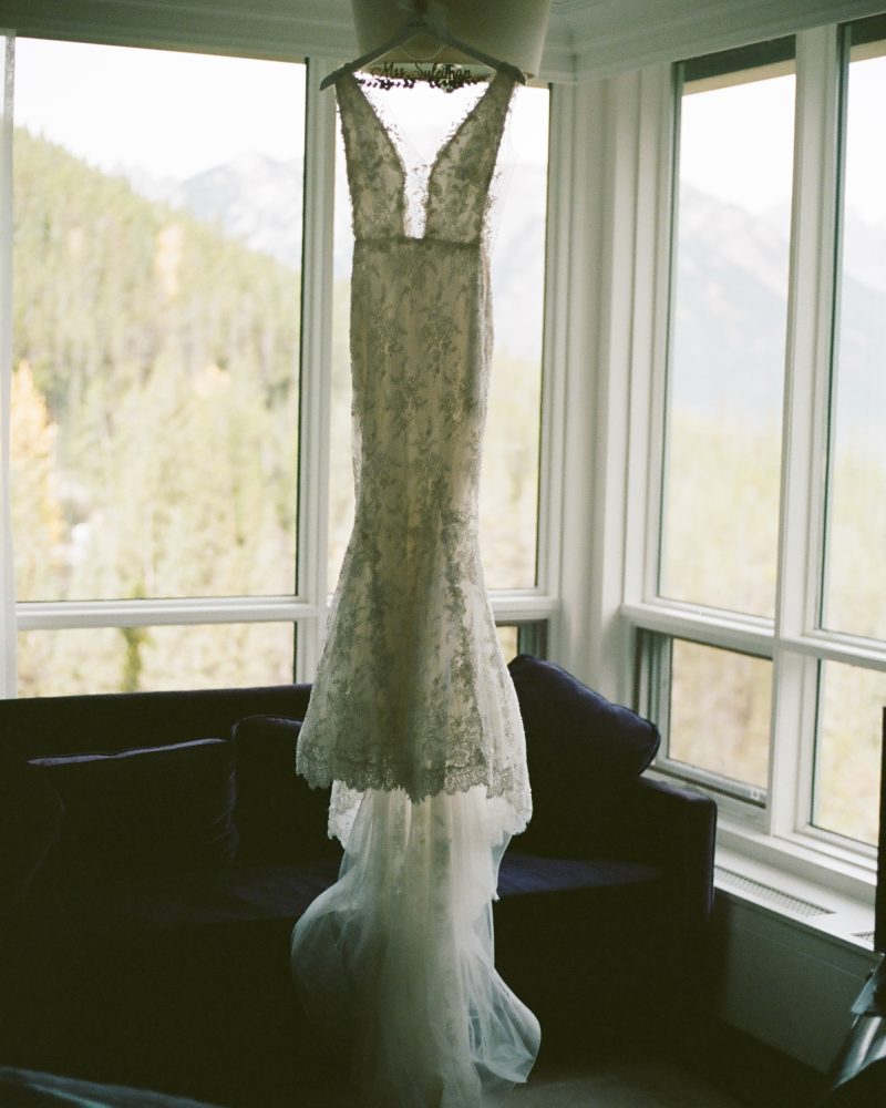 A white lace wedding dress hangs from a light fixture in front of large windows, with a scenic Banff mountain landscape visible outside.