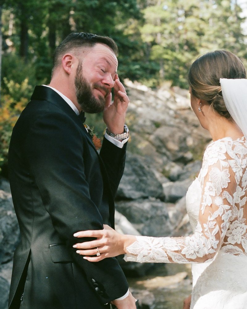 A groom wipes away tears while standing outdoors with his bride, who gently touches his arm. Dressed in wedding attire, they share an intimate elopement surrounded by trees and rocks beside a body of water in Canmore.