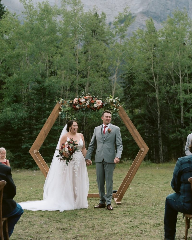 A bride and groom stand hand in hand, smiling under a hexagonal wooden arch decorated with flowers, with a forest and mountain backdrop. Guests are seated outdoors on either side at this intimate Banff elopement.