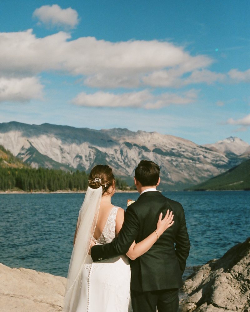 A bride and groom stand arm-in-arm on a rocky shore in Banff, facing a blue lake and distant mountains under a partly cloudy sky. The bride wears a white dress and veil; the groom wears a black suit—perfect for an intimate elopement.