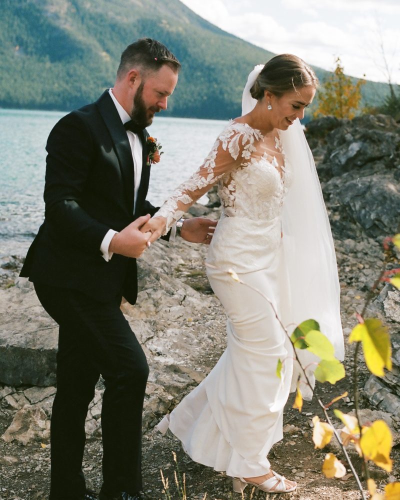 A bride and groom walk hand in hand along a rocky lakeshore in Canmore, with the bride in a white lace gown and veil, and the groom in a black suit. Green mountains and water set the scene for this stunning elopement captured by their photographer.