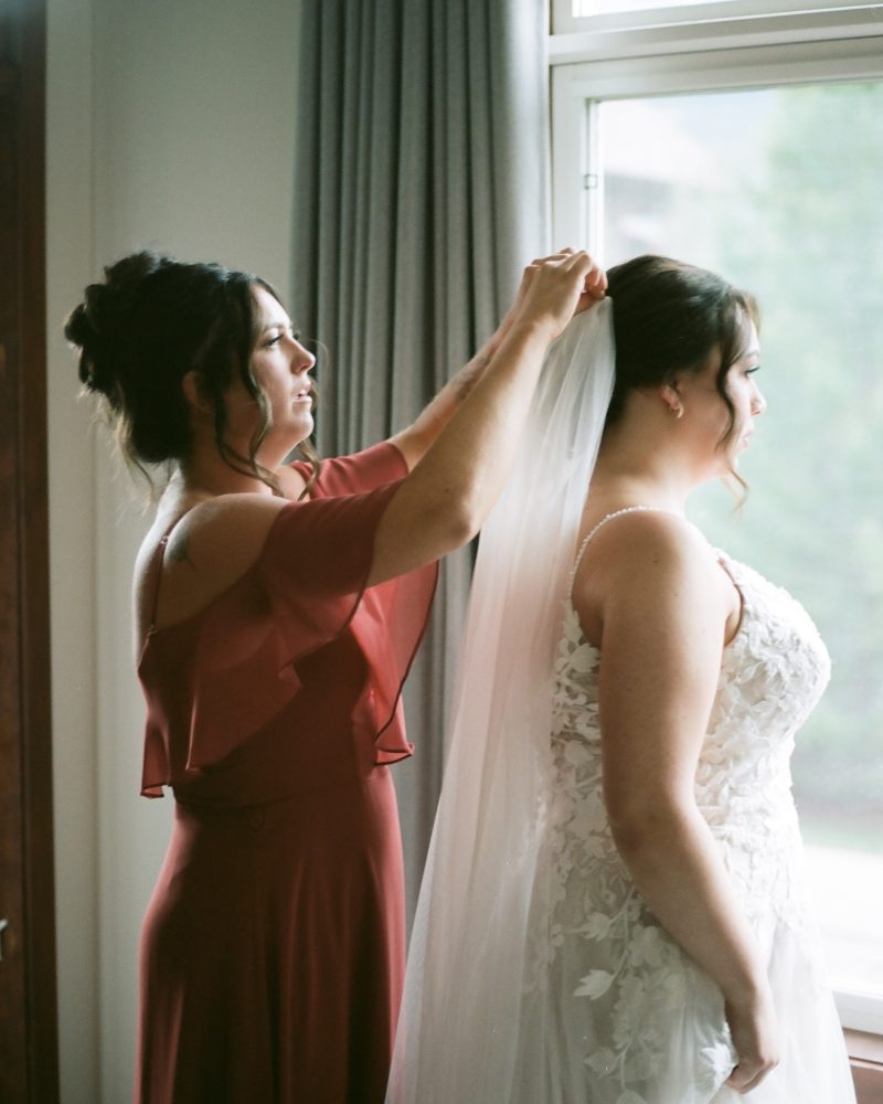 A woman in a red dress helps a bride in a white lace gown adjust her veil near a window with gray curtains, as natural light filters in. Captured by a Canmore photographer, this intimate elopement moment is beautifully soft and serene.