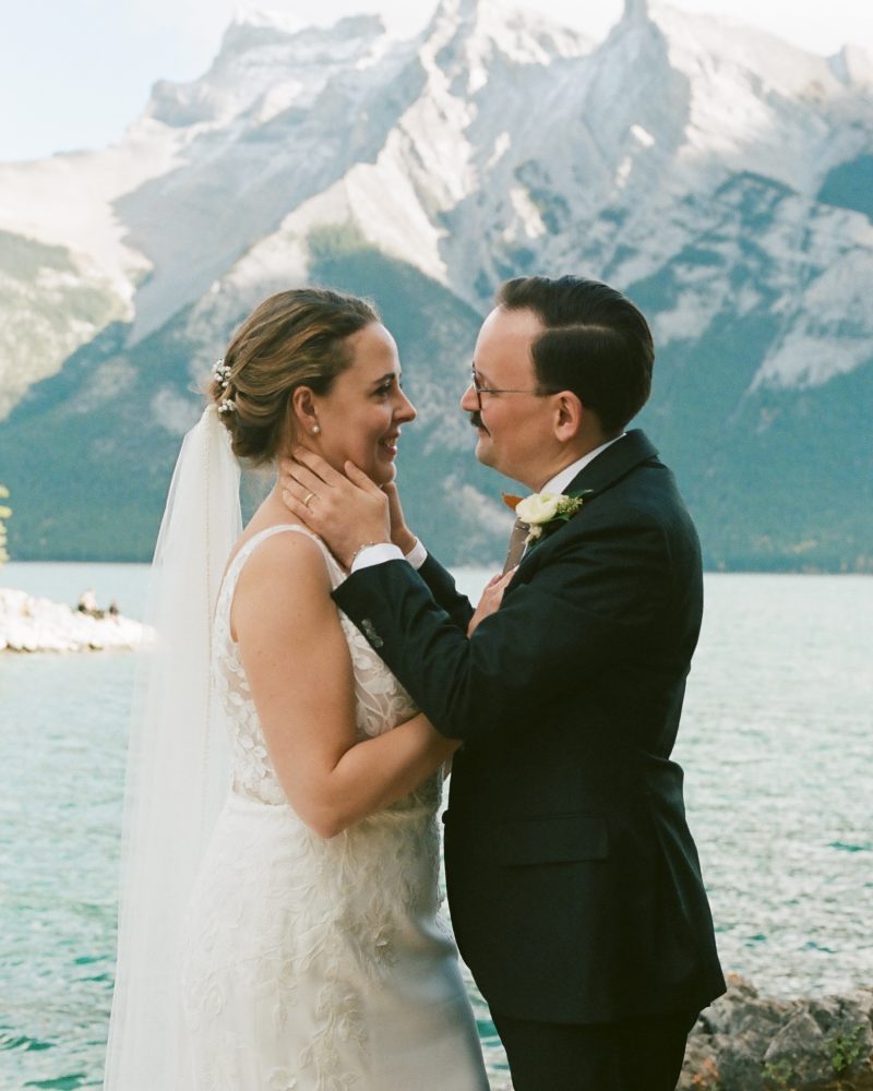 A bride and groom stand by a lake in Banff, smiling and holding each other lovingly. Snow-capped mountains rise in the background under a partly cloudy sky, making their elopement truly unforgettable.