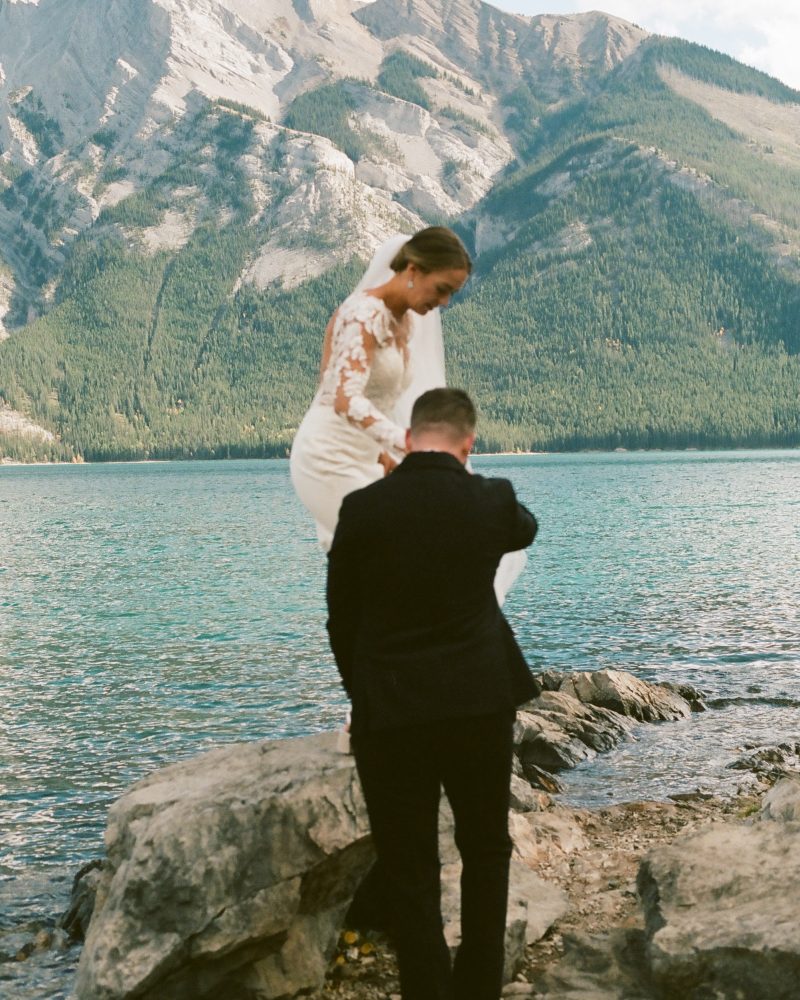A bride in a white dress and veil is helped by a groom in a black suit as she steps onto a rock by a lake near Canmore, with mountains and forest creating a stunning wedding backdrop.