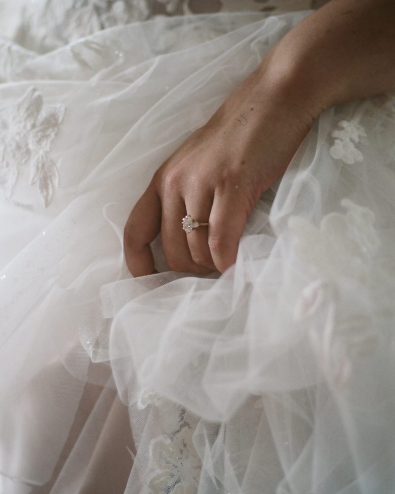 A close-up of a hand resting on a white lace wedding dress during an intimate elopement, showcasing a diamond engagement ring on the ring finger. The delicate fabric and floral details of the dress are beautifully visible.