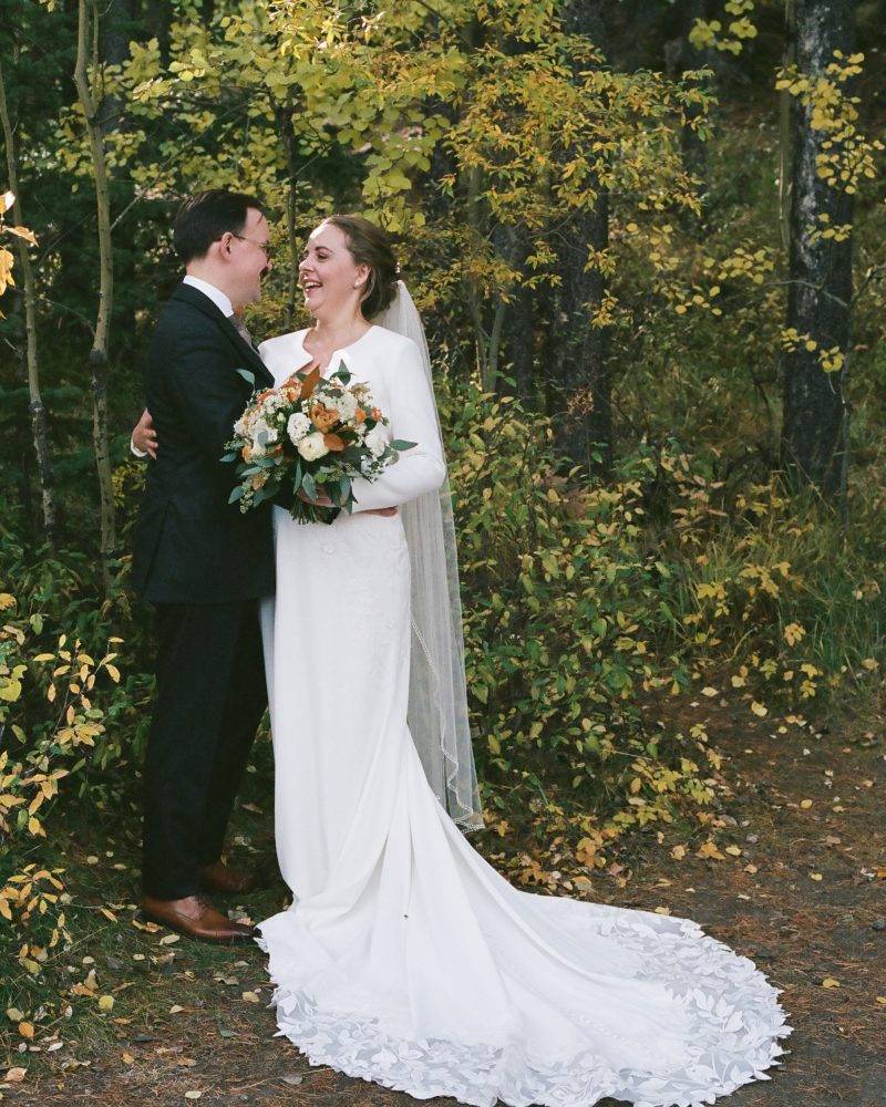 A bride in a long white dress and veil stands smiling with her groom in a dark suit among autumn trees in Banff, holding a bouquet of flowers—perfectly captured by their elopement photographer as they share a joyful moment.