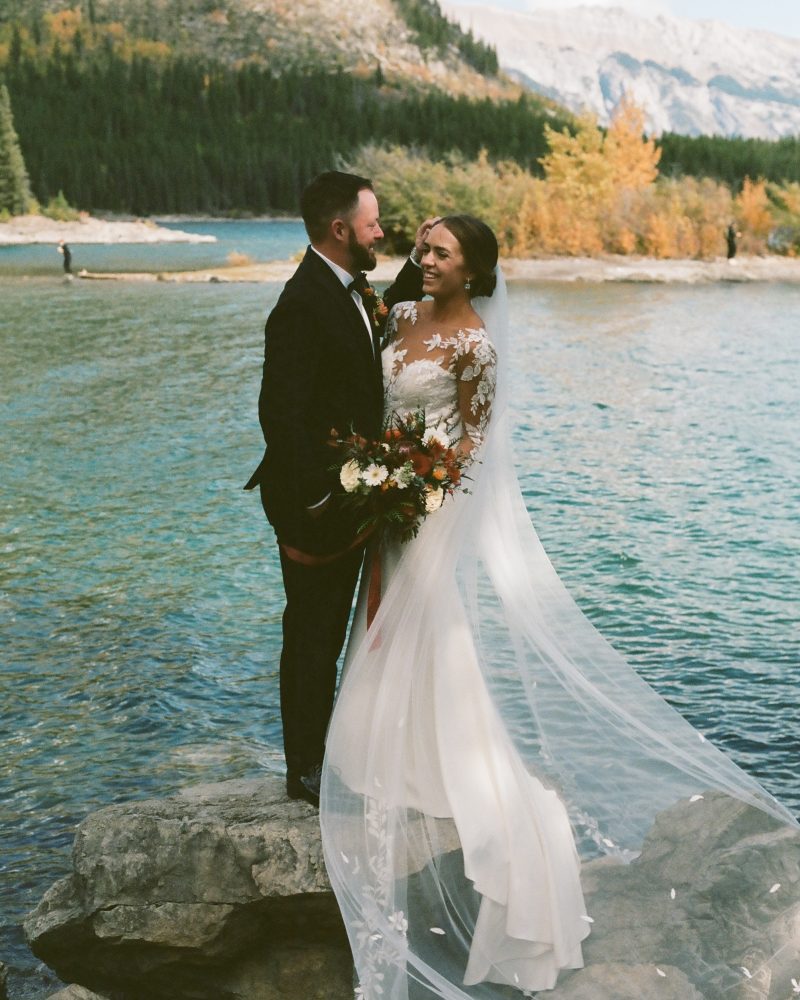 A bride and groom stand on rocks by a lake near Canmore, smiling at each other. The bride wears a long white gown with a veil and holds a bouquet. Forested mountains and blue water stretch out in the background.