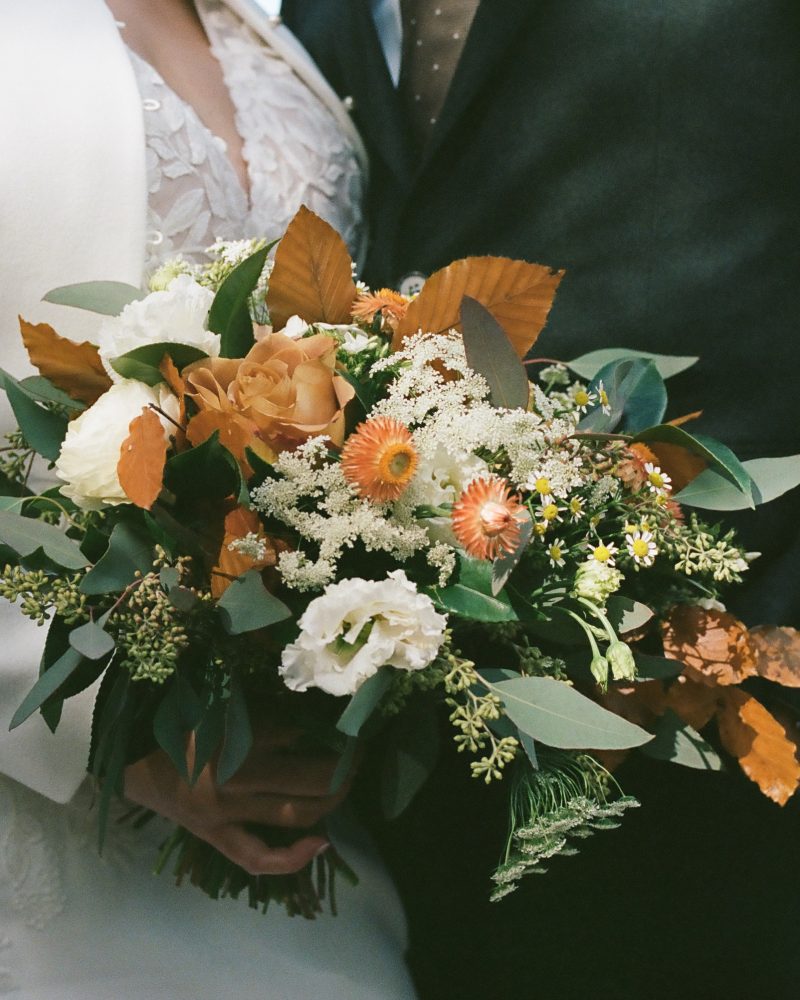 A bride and groom stand side by side outdoors at their Banff elopement; the bride holds a bouquet of white, orange, and green flowers. Their faces are not visible, and the background shows water and blurred scenery.
