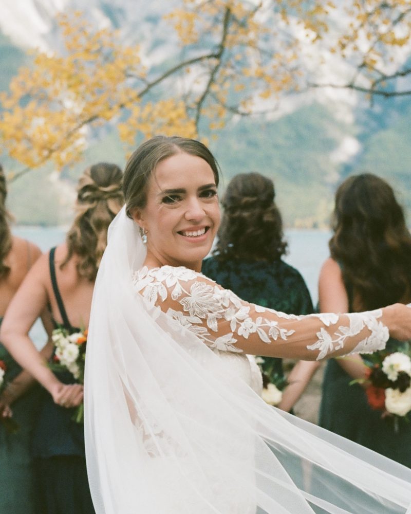 A bride in a white dress smiles at the camera, holding her veil. Three bridesmaids stand behind her, bouquets in hand. Captured by a photographer outdoors near a lake in Banff, with mountains and yellow autumn leaves in the background.