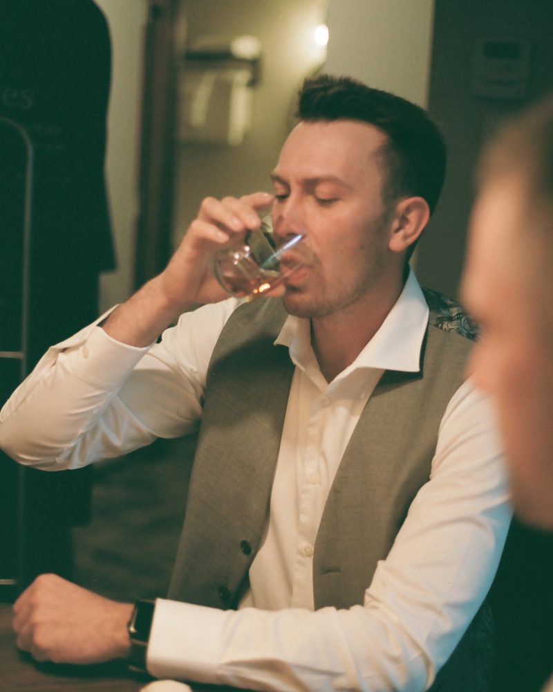 A man in a white shirt and gray vest sits at a table and drinks from a glass. He has short dark hair, wears a smartwatch, and looks like a photographer from Calgary. Another person is partially visible in the foreground indoors.