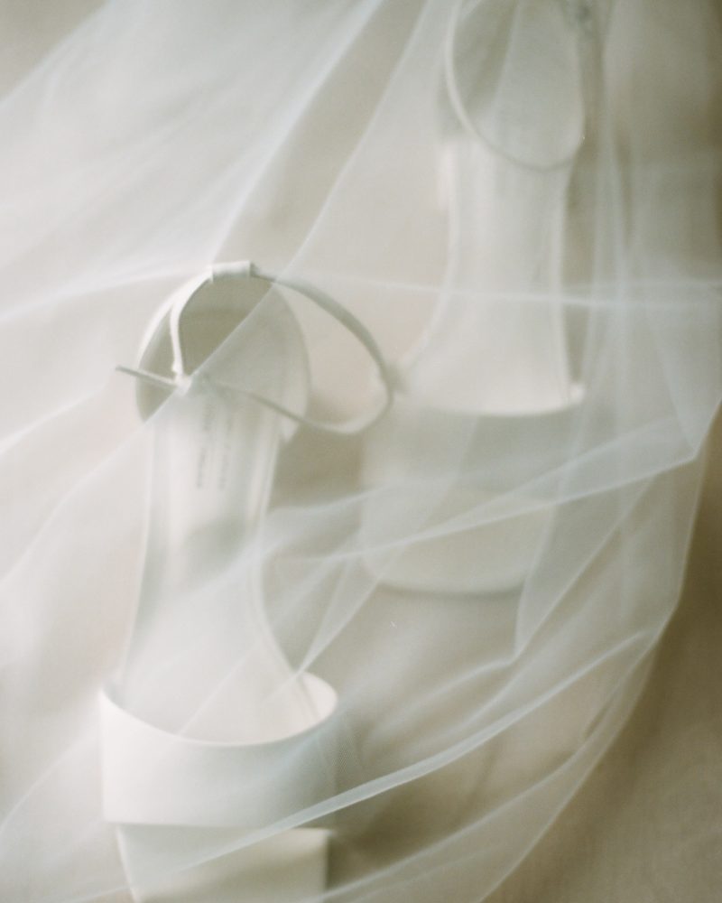 A pair of white open-toe heeled sandals is softly covered by a sheer, delicate white veil on a neutral background, evoking a dreamy and elegant wedding atmosphere inspired by Calgary’s romantic charm.