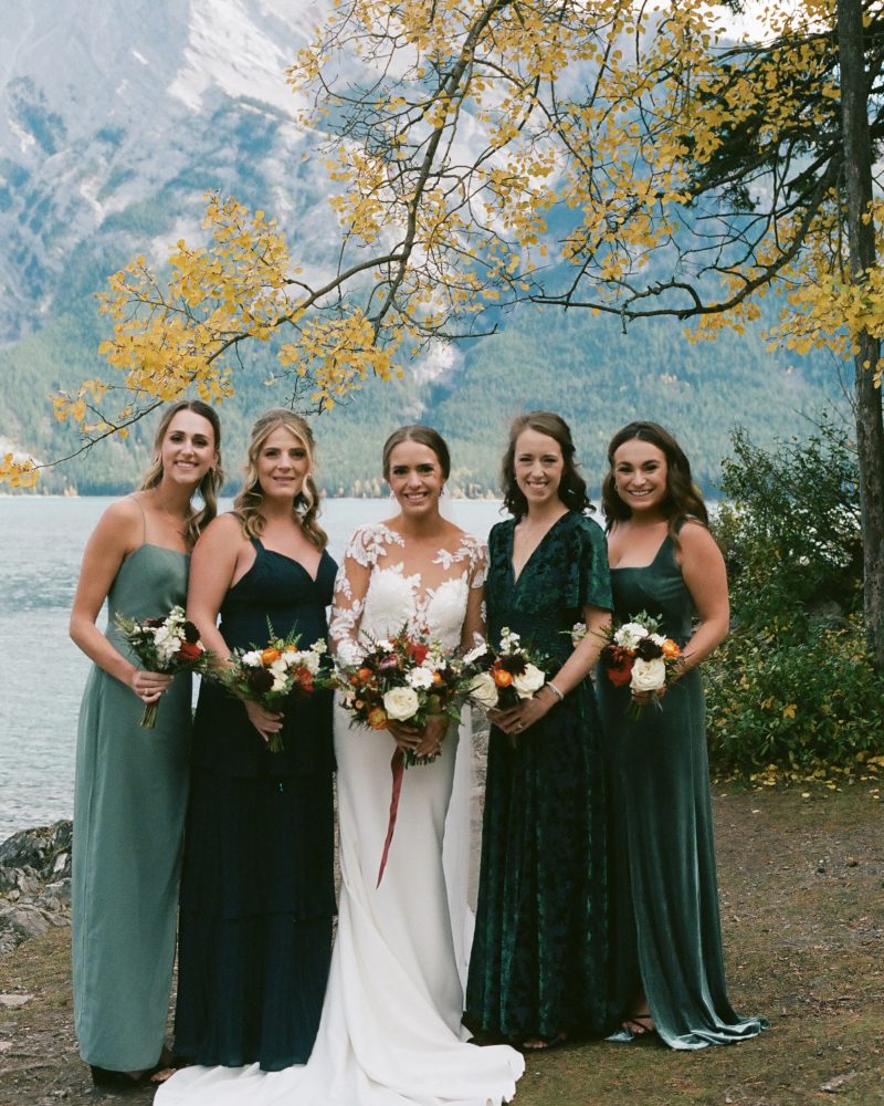 Five women in formal dresses, including one in a white bridal gown, stand outdoors with bouquets. Behind them are autumn trees, a lake, and the mountains near Banff under a cloudy sky—perfect for an intimate elopement.