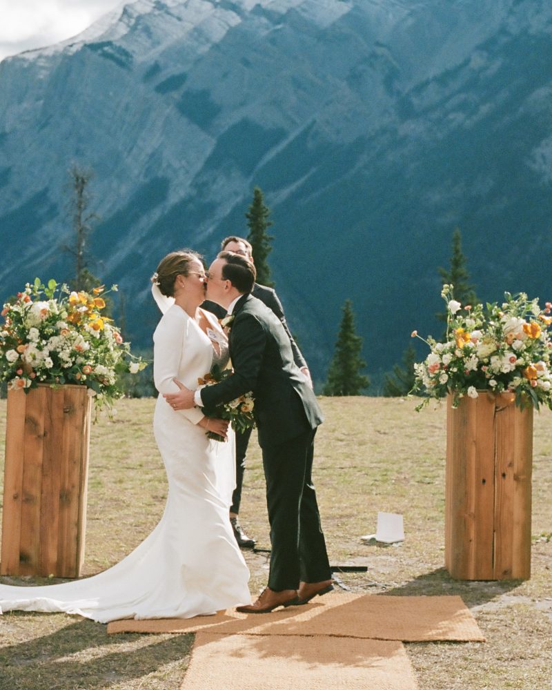 A bride and groom kiss at their Canmore outdoor wedding ceremony with flower arrangements and wooden stands, surrounded by mountains and trees, while guests watch and applaud—perfectly captured by their photographer.