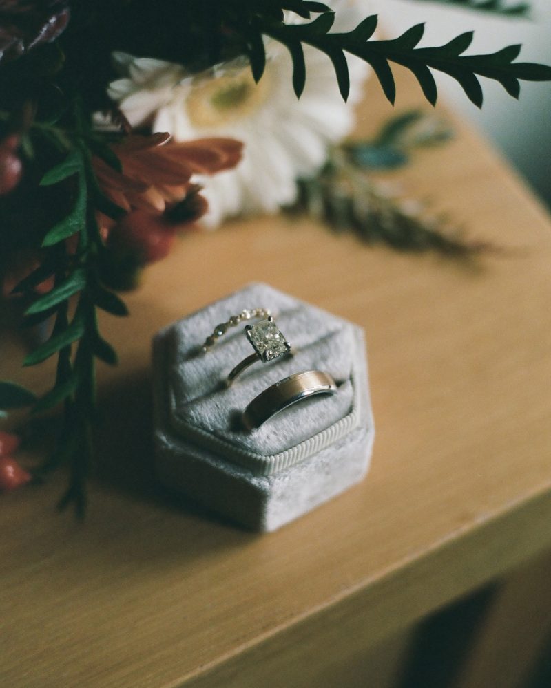 A gold engagement ring and a gold wedding band in a gray velvet hexagonal ring box, placed on a wooden surface next to a bouquet of flowers and greenery—perfect details for an elopement captured by a Banff photographer.