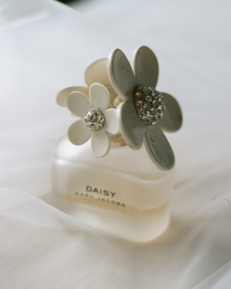 A bottle of Daisy by Marc Jacobs perfume with white flower-shaped cap decorations sits on a soft, white fabric background—perfect for a Canmore wedding or a romantic getaway in Banff.