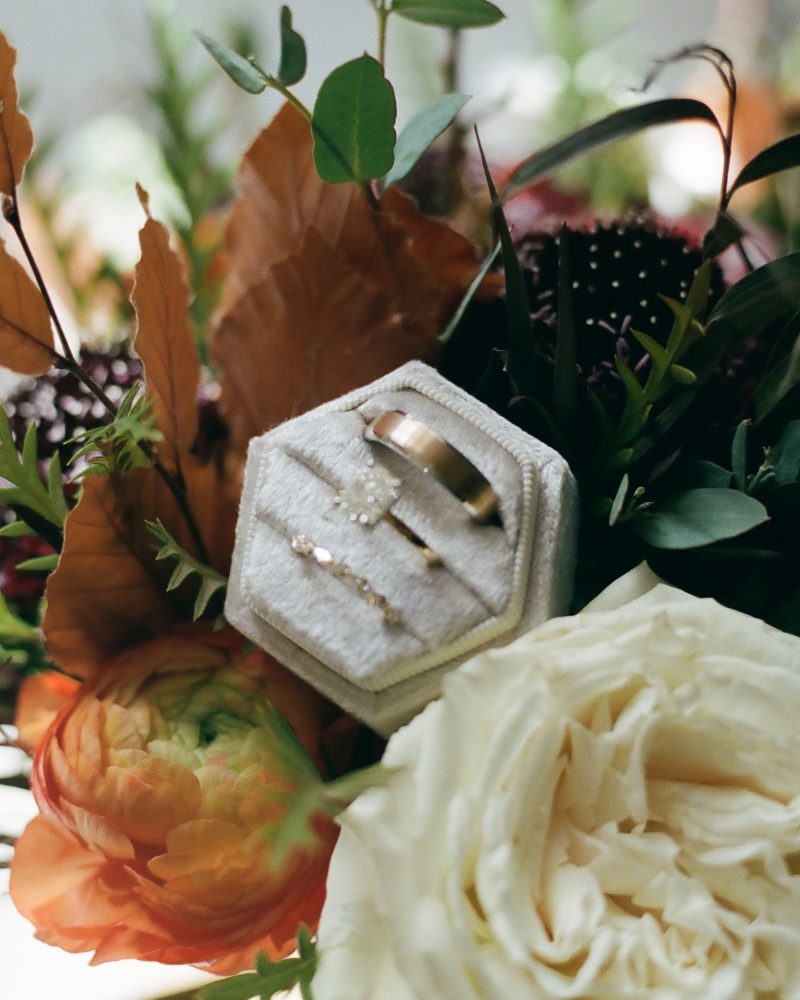 A hexagonal wedding ring box holding three gold rings is nestled among a bouquet of colorful flowers, including white roses, peach ranunculus, and greenery—perfect for a romantic Canmore or Calgary celebration.